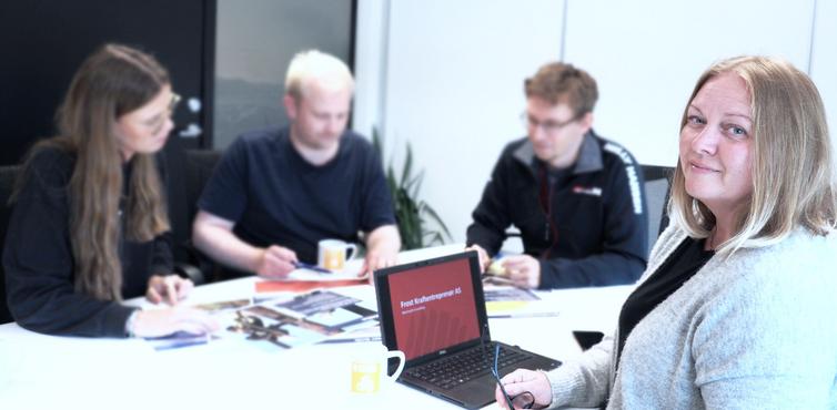 Frost søker nye medarbeidere. Kvinne ser i kamera og prosjektgruppe sitter samlet rundt et bord